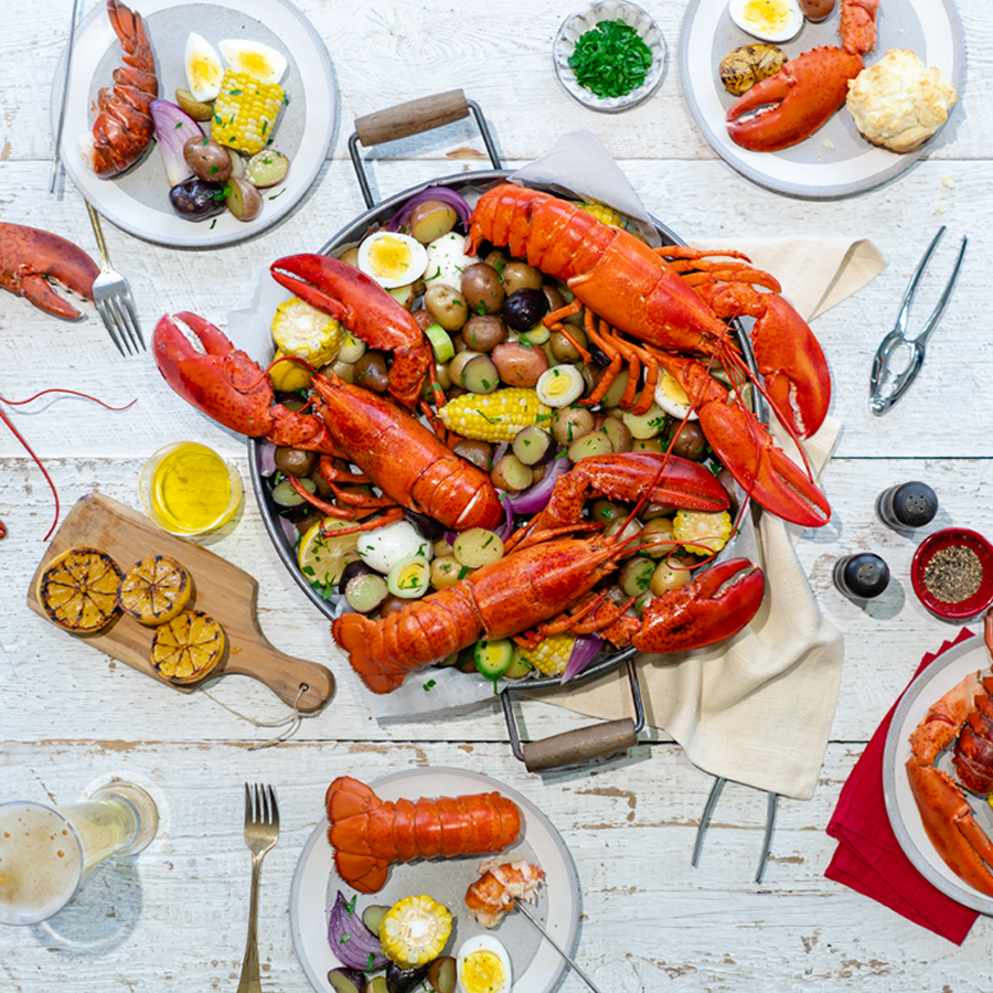 Full Canadian Lobsters are pictured in a large pan with a variety of accompaniments. The pan is on a table set for four with charred lemons, herbs and spices.